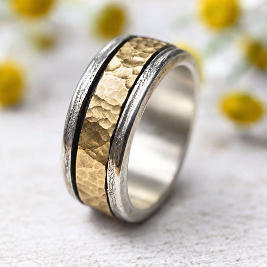 Handgefertigter Spinner-Ring in Silber und 333 Gold mit drehbarem Mittelband, positioniert vor einem zarten Blumenhintergrund aus weißen Blüten mit gelben Punkten.